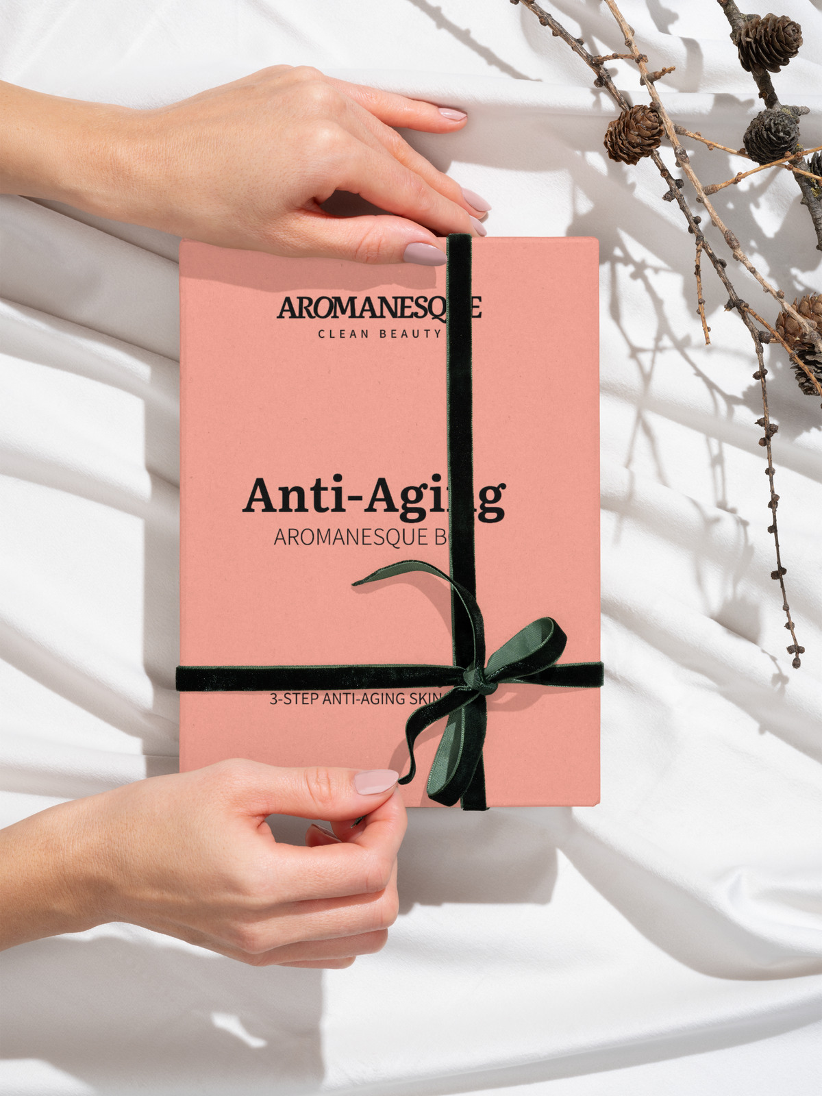A hand holding a pink book titled 'Anti-Aging' by Aromanesque with a decorative bow, placed on a white surface with a background of dry flowers.