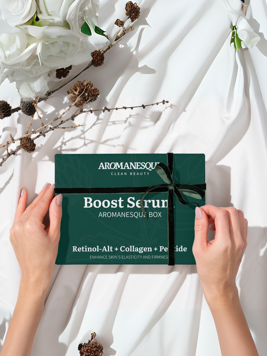 A hand holding a closed green box labeled 'Aromanesque Boost Serum' with a bow on top, presented on a white silky surface with a background of white flowers and some dried flowers.