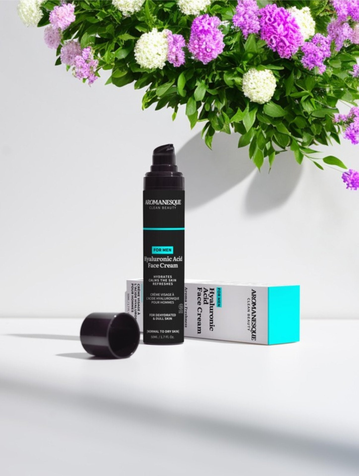 A pump bottle of Aromanesque Hyaluronic Acid Face Cream for Men, with its packaging box and a box of flowers in the background.