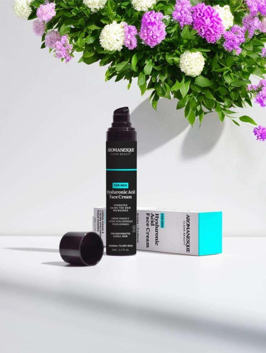 A pump bottle of Aromanesque Hyaluronic Acid Face Cream for Men, with its packaging box and a box of flowers in the background.